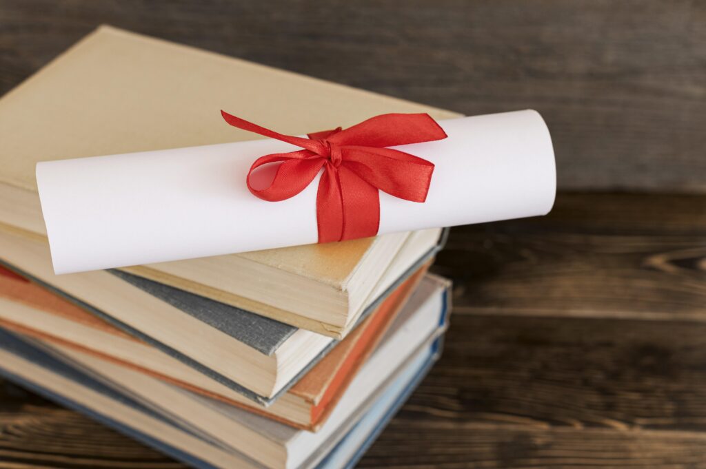 image shows a stack of books on a wooden surface with a diploma tied with a red ribbon resting on top